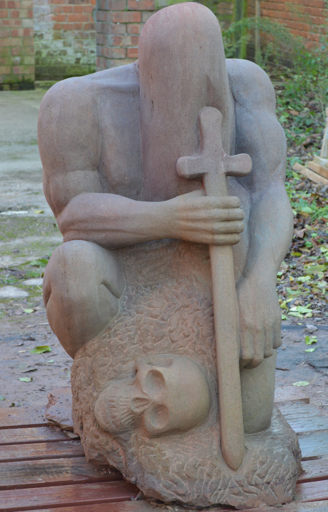 Deborah Harrison sculpture The Guardian, a monumental one-off sandstone figure kneeling with bowed head, gripping a sword planted into the ground. Carved from warm Forest of Dean sandstone, the faceless guardian form conveys strength, sacrifice, and timeless watchfulness, with a skull motif at its base adding symbolism of mortality and protection.