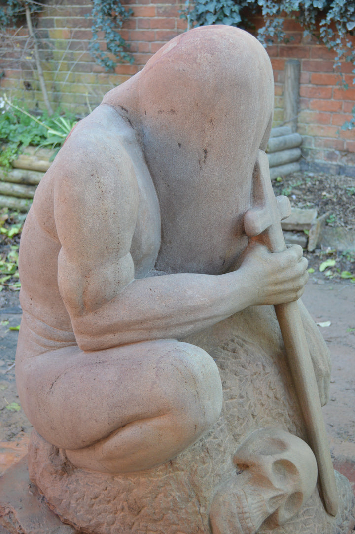 Deborah Harrison sculpture The Guardian, a monumental one-off sandstone figure kneeling with bowed head, gripping a sword planted into the ground. Carved from warm Forest of Dean sandstone, the faceless guardian form conveys strength, sacrifice, and timeless watchfulness, with a skull motif at its base adding symbolism of mortality and protection.