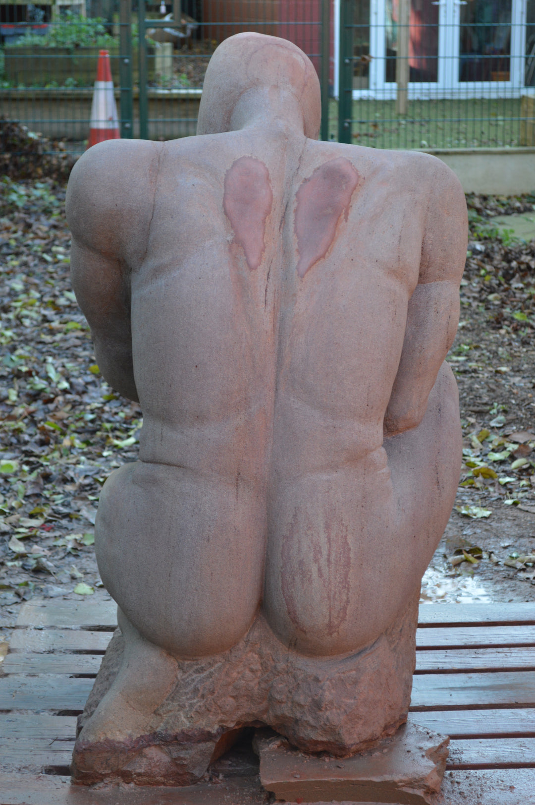 Deborah Harrison sculpture The Guardian, a monumental one-off sandstone figure kneeling with bowed head, gripping a sword planted into the ground. Carved from warm Forest of Dean sandstone, the faceless guardian form conveys strength, sacrifice, and timeless watchfulness, with a skull motif at its base adding symbolism of mortality and protection.