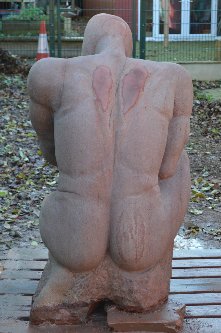 Deborah Harrison sculpture The Guardian, a monumental one-off sandstone figure kneeling with bowed head, gripping a sword planted into the ground. Carved from warm Forest of Dean sandstone, the faceless guardian form conveys strength, sacrifice, and timeless watchfulness, with a skull motif at its base adding symbolism of mortality and protection.