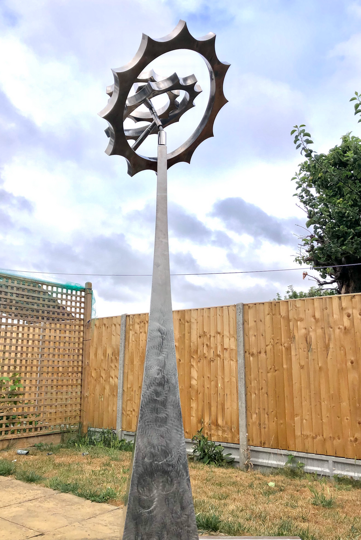 Kinetic sculpture by Martin Jarvis featuring intersecting stainless steel rings with sculpted, gear-like outer edges, designed to rotate on multiple axes atop a tall, tapered plinth. The polished metal reflects the sky and surroundings, enhancing the illusion of perpetual motion and balance.