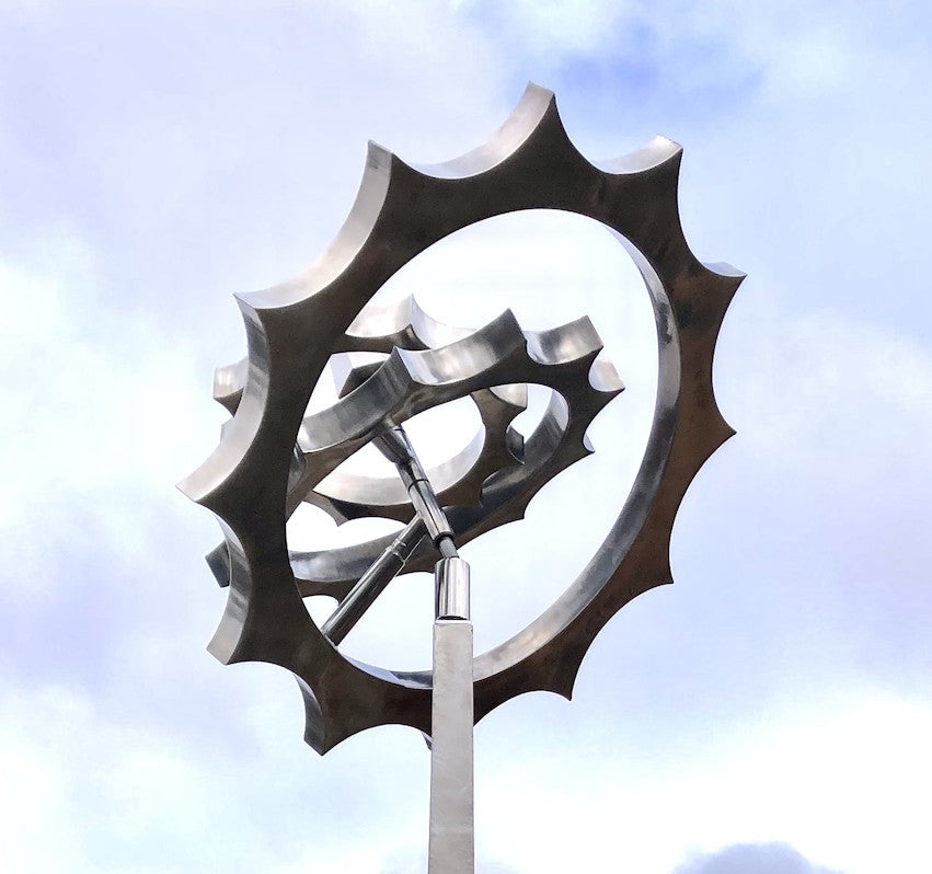 Kinetic sculpture by Martin Jarvis featuring intersecting stainless steel rings with sculpted, gear-like outer edges, designed to rotate on multiple axes atop a tall, tapered plinth. The polished metal reflects the sky and surroundings, enhancing the illusion of perpetual motion and balance.