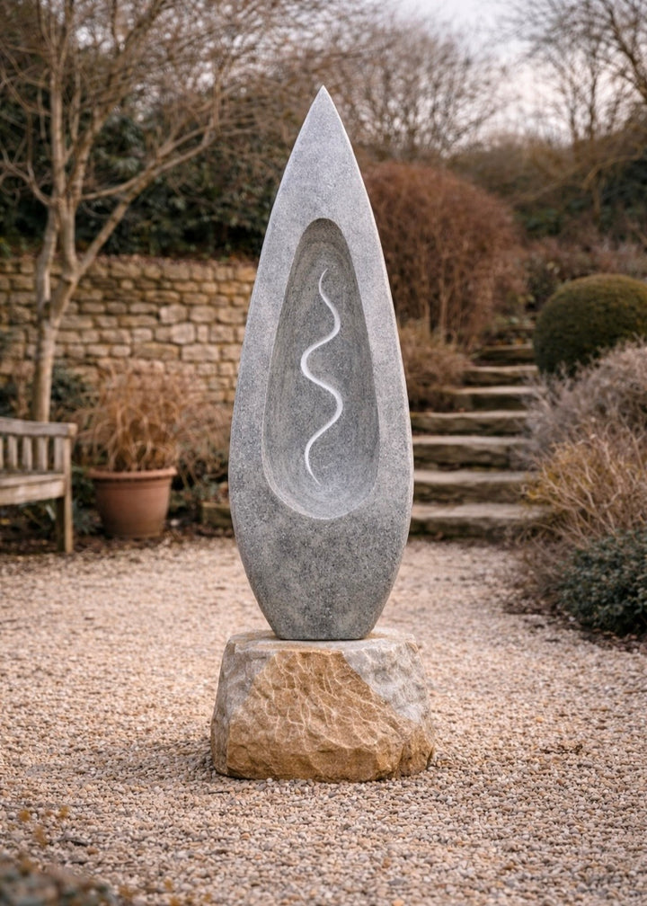 Tall grey Ancaster stone sculpture by Martin Cash with carved hollow centre and flowing white vein motif, mounted on a natural stone base in a landscaped garden setting.