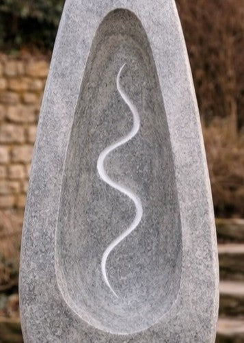 Tall grey Ancaster stone sculpture by Martin Cash with carved hollow centre and flowing white vein motif, mounted on a natural stone base in a landscaped garden setting.