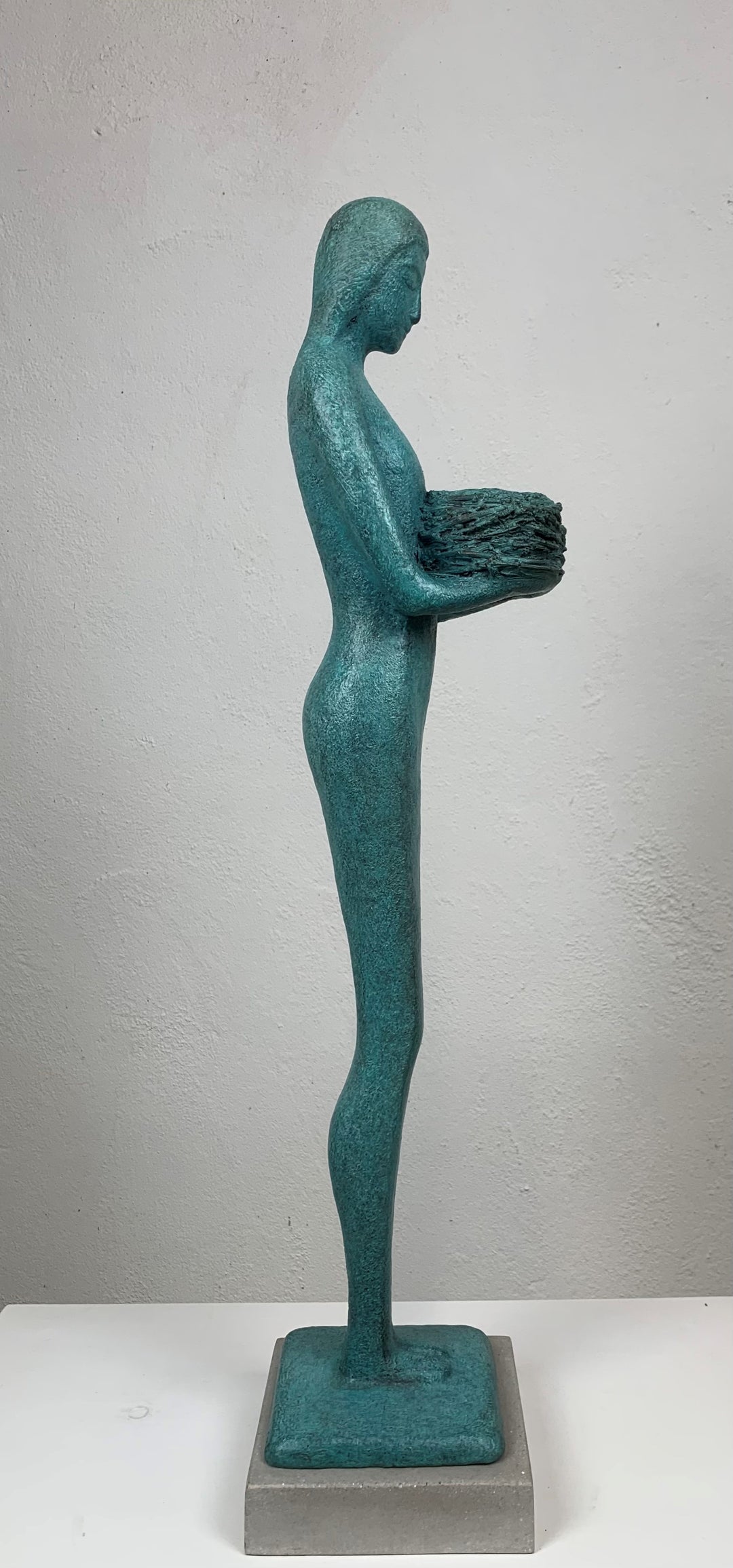 Elegant teal bronze sculpture of a standing female figure in profile, holding a textured nest-like form close to her chest.