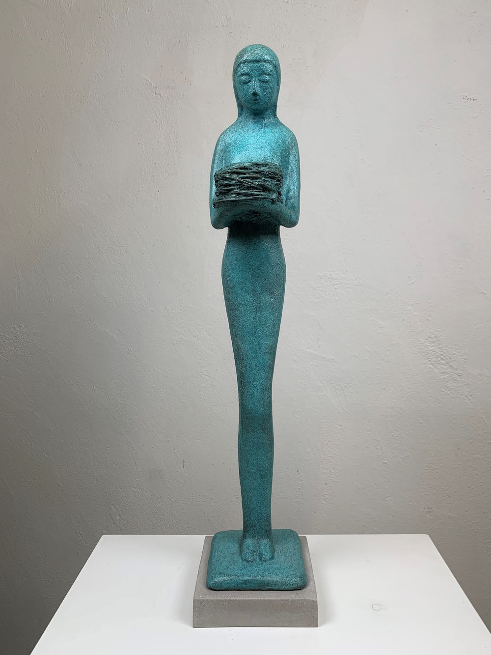 Elegant teal bronze sculpture of a standing female figure in profile, holding a textured nest-like form close to her chest.