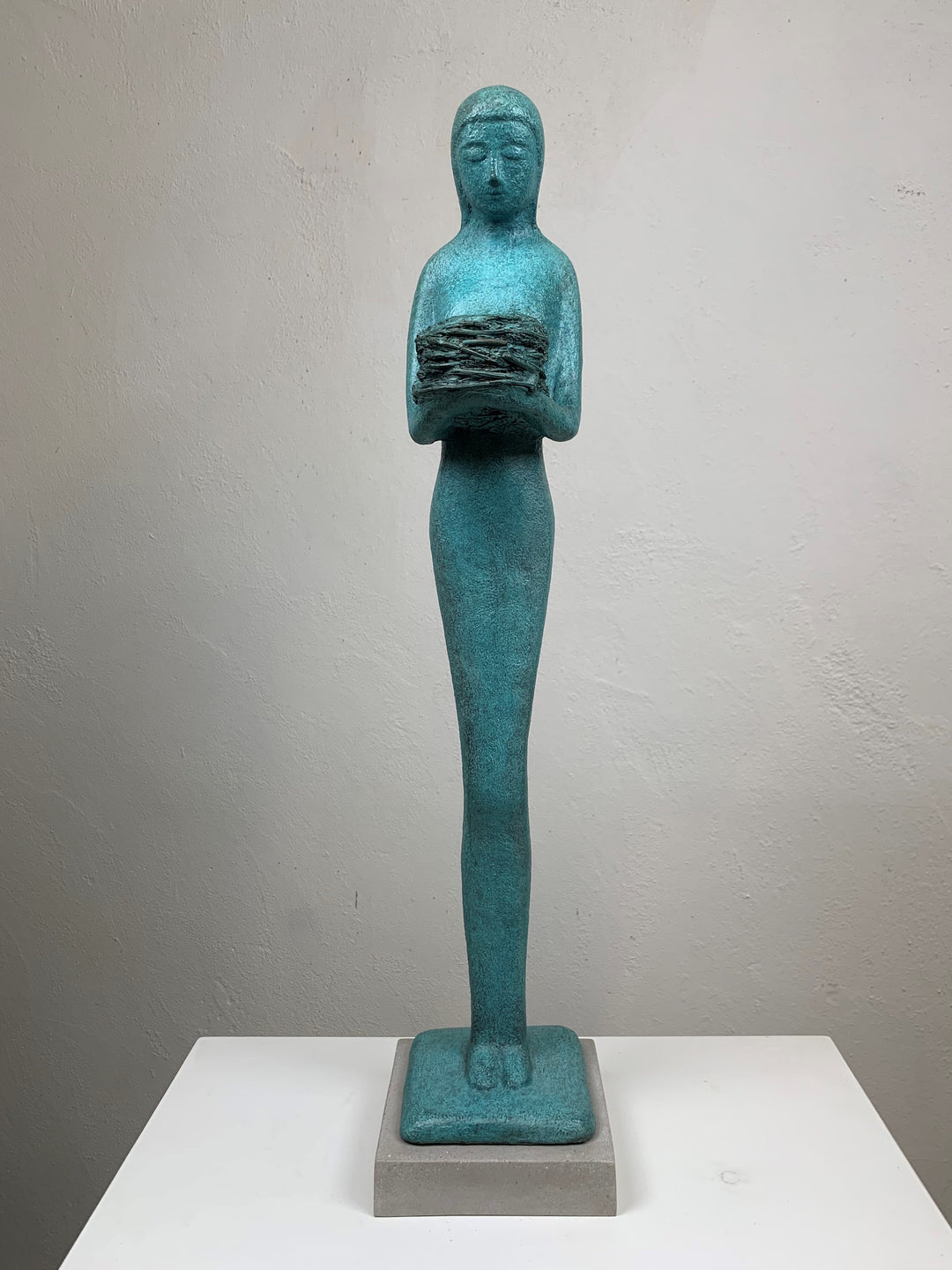Elegant teal bronze sculpture of a standing female figure in profile, holding a textured nest-like form close to her chest.