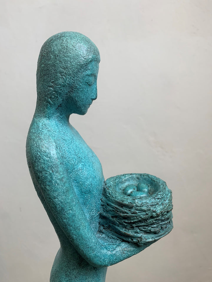 Elegant teal bronze sculpture of a standing female figure in profile, holding a textured nest-like form close to her chest.
