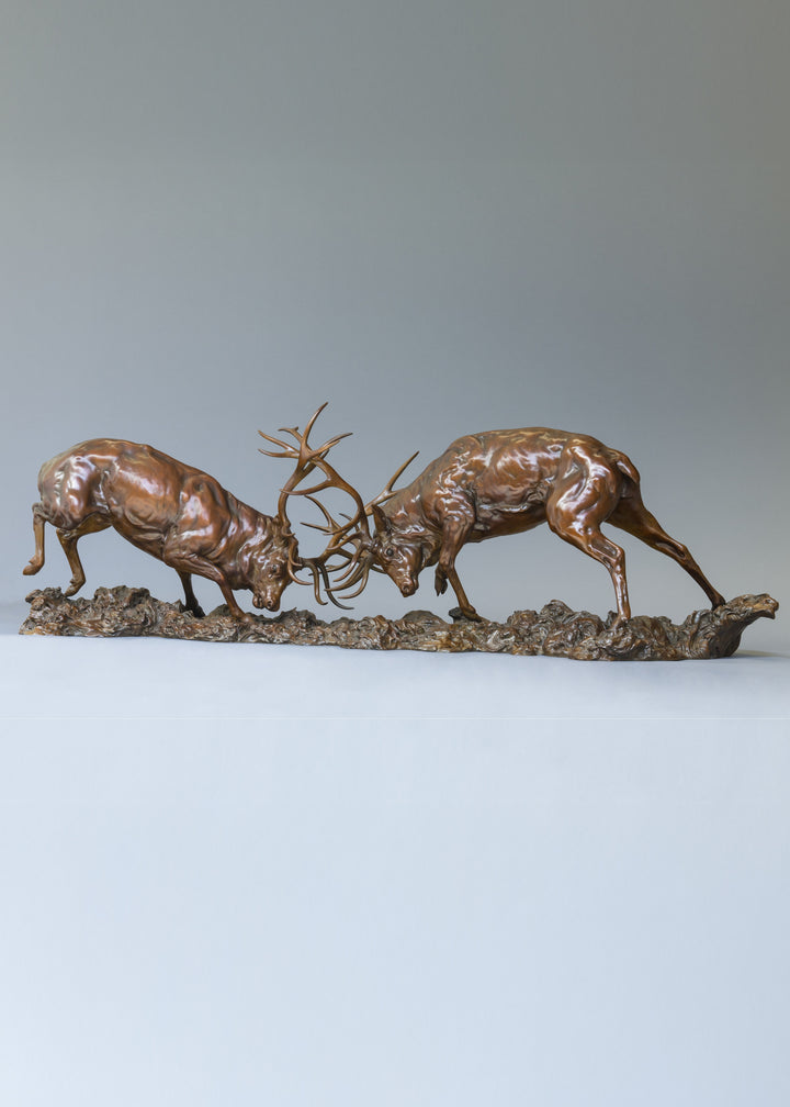 Bronze sculpture of two red deer stags in mid-rut, antlers locked in a dynamic head-to-head clash, highly detailed musculature and patina, mounted on a textured bronze base.