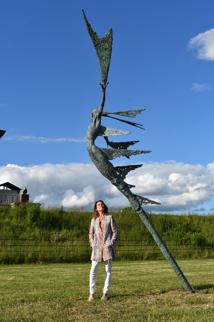 Monumental bronze sculpture of Theia, Greek goddess of light, portrayed in an elegant, elongated form with flowing limbs and wing-like extensions. Her textured surface and dynamic pose evoke movement and radiance, with light-catching shards projecting from her figure against open sky and landscape.