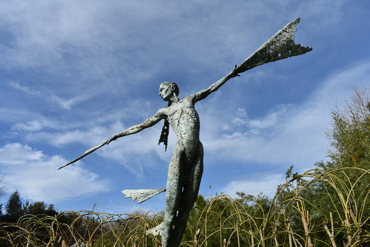 Monumental bronze sculpture of Theia, Greek goddess of light, portrayed in an elegant, elongated form with flowing limbs and wing-like extensions. Her textured surface and dynamic pose evoke movement and radiance, with light-catching shards projecting from her figure against open sky and landscape.