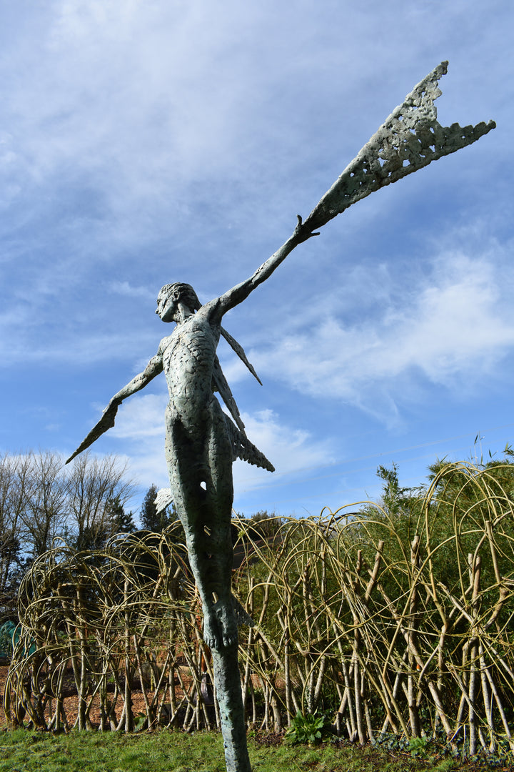 Monumental bronze sculpture of Theia, Greek goddess of light, portrayed in an elegant, elongated form with flowing limbs and wing-like extensions. Her textured surface and dynamic pose evoke movement and radiance, with light-catching shards projecting from her figure against open sky and landscape.