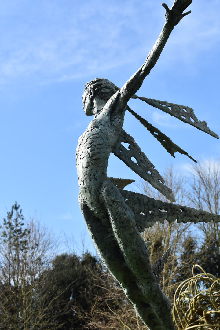 Monumental bronze sculpture of Theia, Greek goddess of light, portrayed in an elegant, elongated form with flowing limbs and wing-like extensions. Her textured surface and dynamic pose evoke movement and radiance, with light-catching shards projecting from her figure against open sky and landscape.