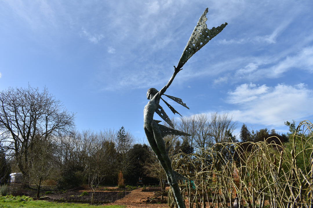 Monumental bronze sculpture of Theia, Greek goddess of light, portrayed in an elegant, elongated form with flowing limbs and wing-like extensions. Her textured surface and dynamic pose evoke movement and radiance, with light-catching shards projecting from her figure against open sky and landscape.