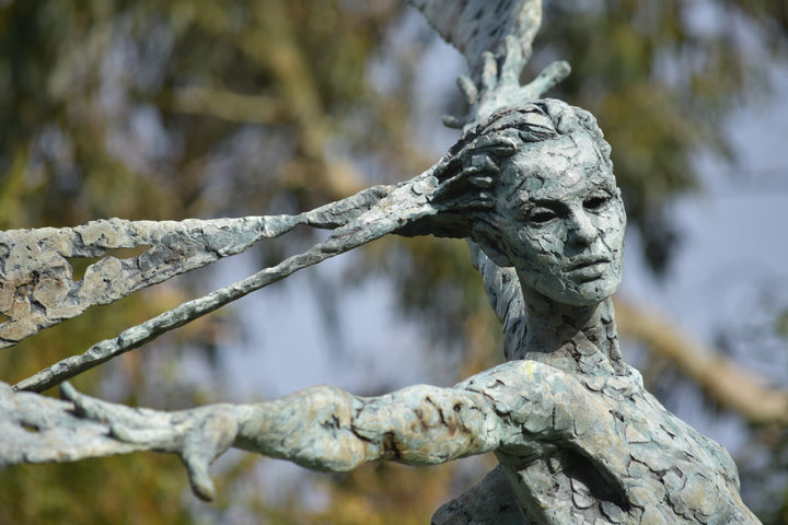 Monumental bronze sculpture of Theia, Greek goddess of light, portrayed in an elegant, elongated form with flowing limbs and wing-like extensions. Her textured surface and dynamic pose evoke movement and radiance, with light-catching shards projecting from her figure against open sky and landscape.