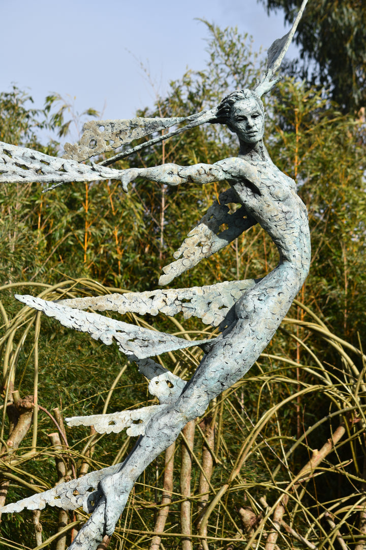 Monumental bronze sculpture of Theia, Greek goddess of light, portrayed in an elegant, elongated form with flowing limbs and wing-like extensions. Her textured surface and dynamic pose evoke movement and radiance, with light-catching shards projecting from her figure against open sky and landscape.