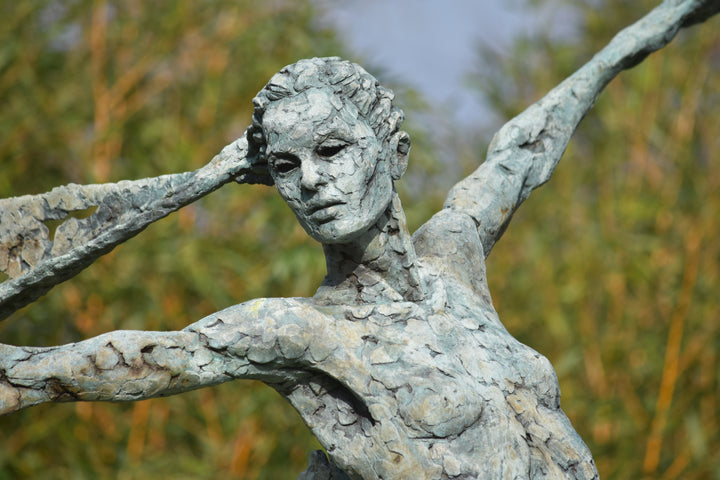 Monumental bronze sculpture of Theia, Greek goddess of light, portrayed in an elegant, elongated form with flowing limbs and wing-like extensions. Her textured surface and dynamic pose evoke movement and radiance, with light-catching shards projecting from her figure against open sky and landscape.