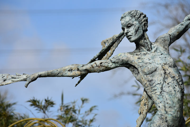 Monumental bronze sculpture of Theia, Greek goddess of light, portrayed in an elegant, elongated form with flowing limbs and wing-like extensions. Her textured surface and dynamic pose evoke movement and radiance, with light-catching shards projecting from her figure against open sky and landscape.