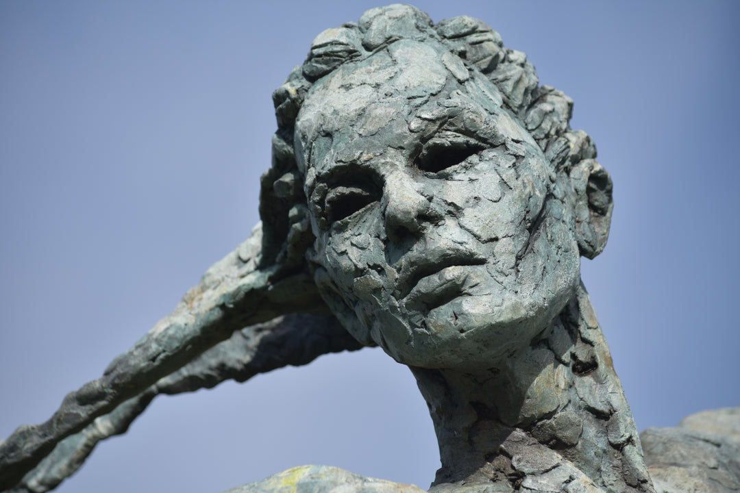Monumental bronze sculpture of Theia, Greek goddess of light, portrayed in an elegant, elongated form with flowing limbs and wing-like extensions. Her textured surface and dynamic pose evoke movement and radiance, with light-catching shards projecting from her figure against open sky and landscape.