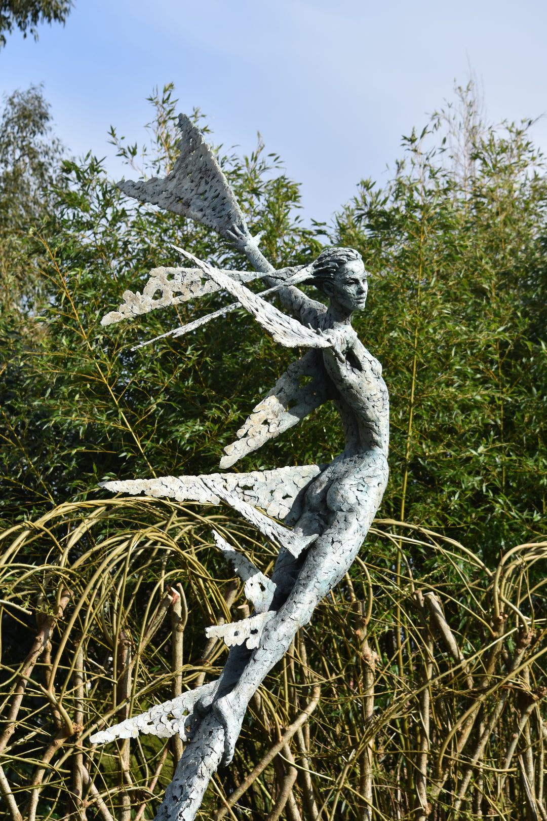 Monumental bronze sculpture of Theia, Greek goddess of light, portrayed in an elegant, elongated form with flowing limbs and wing-like extensions. Her textured surface and dynamic pose evoke movement and radiance, with light-catching shards projecting from her figure against open sky and landscape.