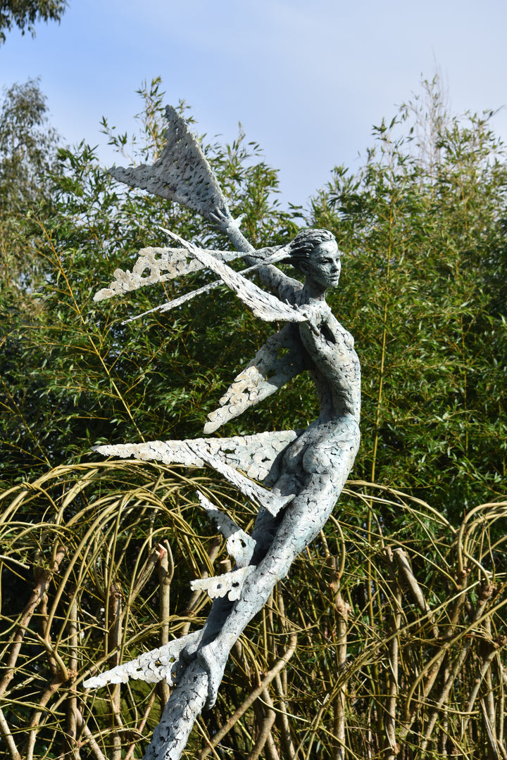 Monumental bronze sculpture of Theia, Greek goddess of light, portrayed in an elegant, elongated form with flowing limbs and wing-like extensions. Her textured surface and dynamic pose evoke movement and radiance, with light-catching shards projecting from her figure against open sky and landscape.