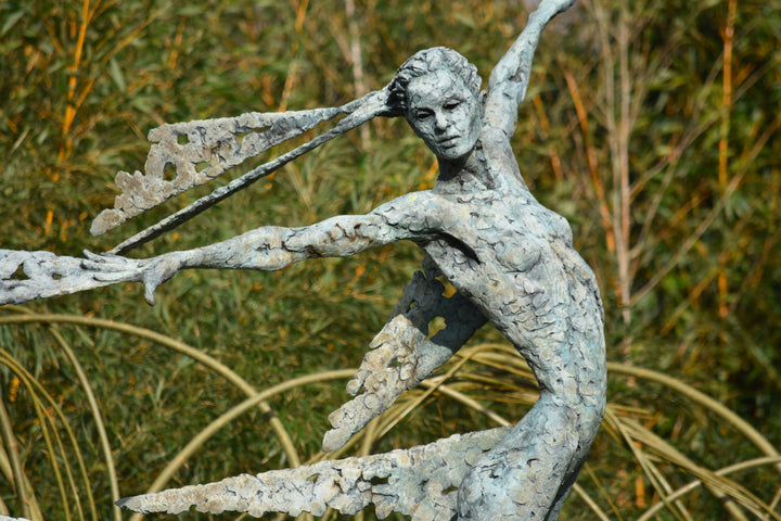 Monumental bronze sculpture of Theia, Greek goddess of light, portrayed in an elegant, elongated form with flowing limbs and wing-like extensions. Her textured surface and dynamic pose evoke movement and radiance, with light-catching shards projecting from her figure against open sky and landscape.