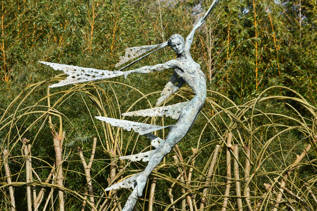 Monumental bronze sculpture of Theia, Greek goddess of light, portrayed in an elegant, elongated form with flowing limbs and wing-like extensions. Her textured surface and dynamic pose evoke movement and radiance, with light-catching shards projecting from her figure against open sky and landscape.