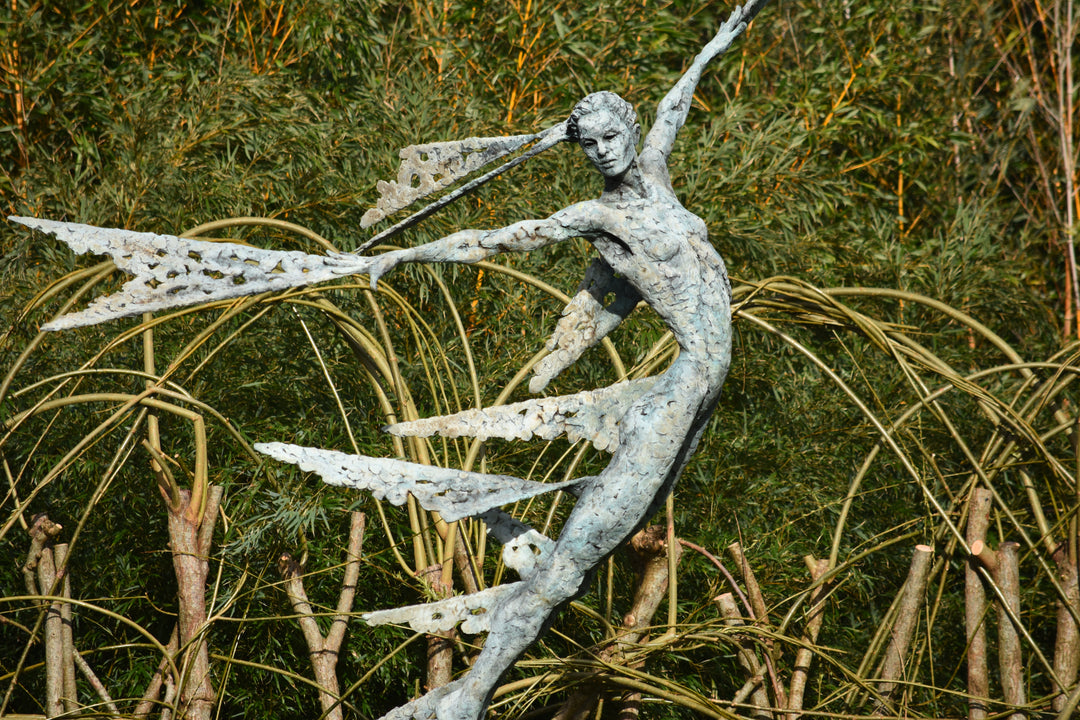 Monumental bronze sculpture of Theia, Greek goddess of light, portrayed in an elegant, elongated form with flowing limbs and wing-like extensions. Her textured surface and dynamic pose evoke movement and radiance, with light-catching shards projecting from her figure against open sky and landscape.