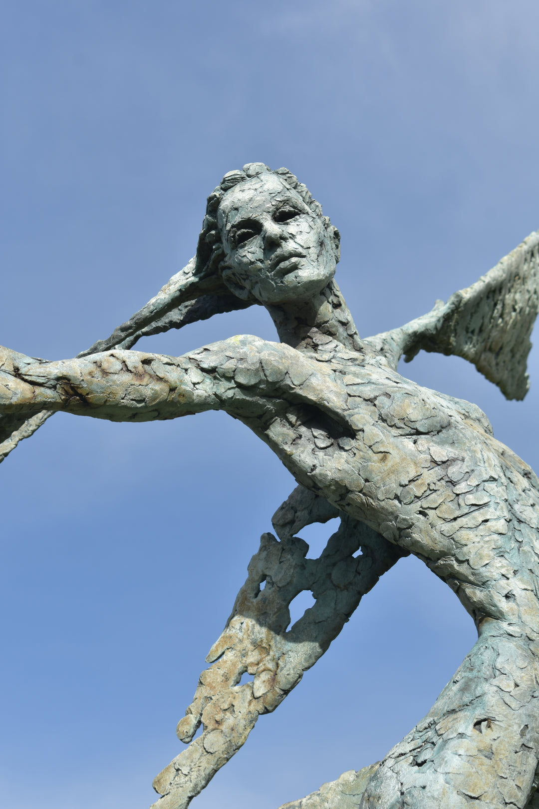 Monumental bronze sculpture of Theia, Greek goddess of light, portrayed in an elegant, elongated form with flowing limbs and wing-like extensions. Her textured surface and dynamic pose evoke movement and radiance, with light-catching shards projecting from her figure against open sky and landscape.