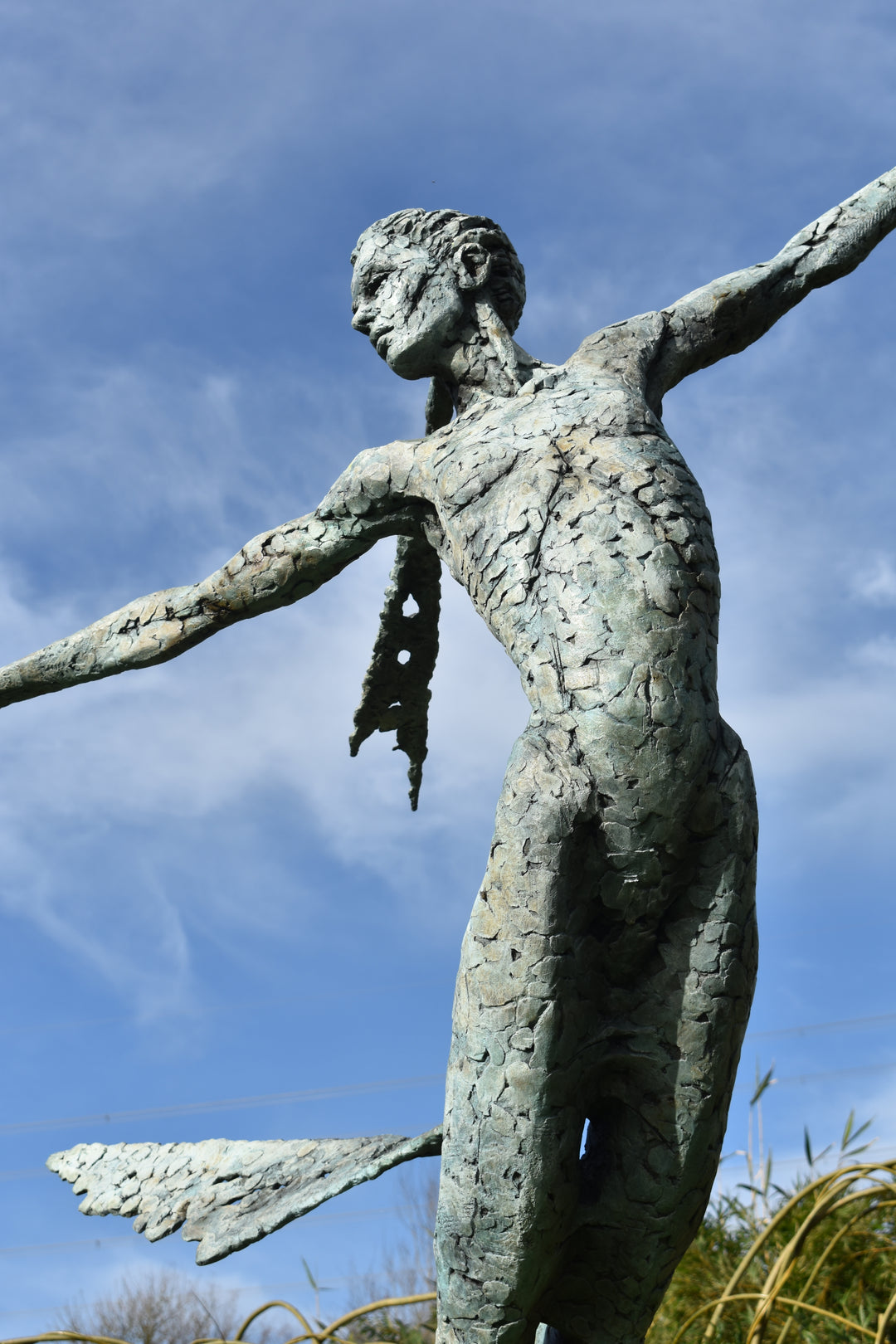 Monumental bronze sculpture of Theia, Greek goddess of light, portrayed in an elegant, elongated form with flowing limbs and wing-like extensions. Her textured surface and dynamic pose evoke movement and radiance, with light-catching shards projecting from her figure against open sky and landscape.