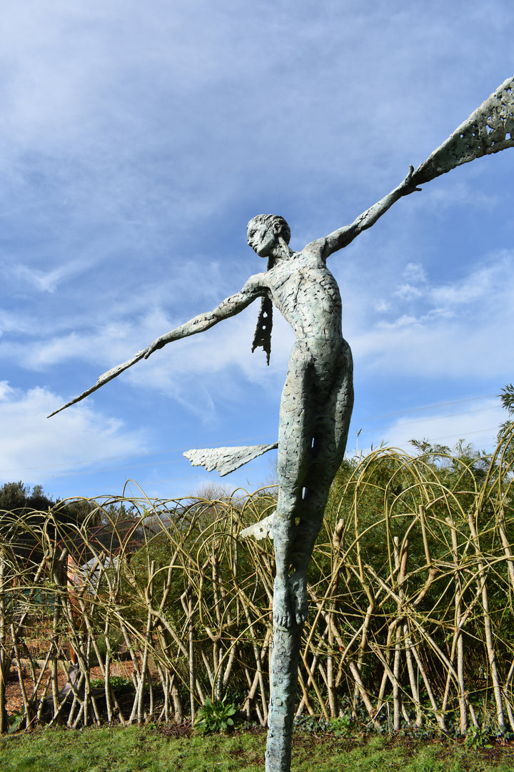 Monumental bronze sculpture of Theia, Greek goddess of light, portrayed in an elegant, elongated form with flowing limbs and wing-like extensions. Her textured surface and dynamic pose evoke movement and radiance, with light-catching shards projecting from her figure against open sky and landscape.