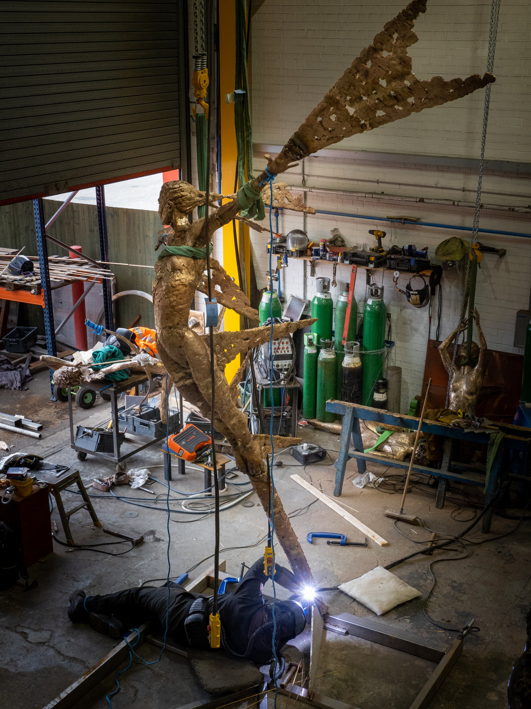 Monumental bronze sculpture of Theia, Greek goddess of light, portrayed in an elegant, elongated form with flowing limbs and wing-like extensions. Her textured surface and dynamic pose evoke movement and radiance, with light-catching shards projecting from her figure against open sky and landscape.