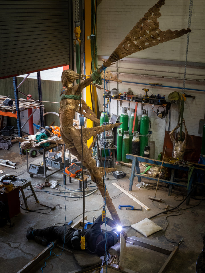 Monumental bronze sculpture of Theia, Greek goddess of light, portrayed in an elegant, elongated form with flowing limbs and wing-like extensions. Her textured surface and dynamic pose evoke movement and radiance, with light-catching shards projecting from her figure against open sky and landscape.