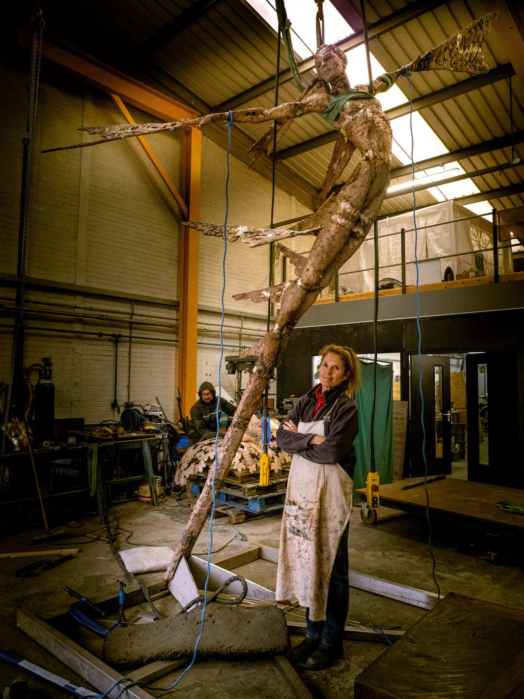 Monumental bronze sculpture of Theia, Greek goddess of light, portrayed in an elegant, elongated form with flowing limbs and wing-like extensions. Her textured surface and dynamic pose evoke movement and radiance, with light-catching shards projecting from her figure against open sky and landscape.