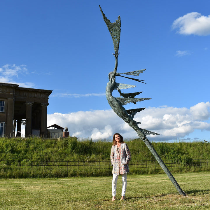 Monumental bronze sculpture of Theia, Greek goddess of light, portrayed in an elegant, elongated form with flowing limbs and wing-like extensions. Her textured surface and dynamic pose evoke movement and radiance, with light-catching shards projecting from her figure against open sky and landscape.