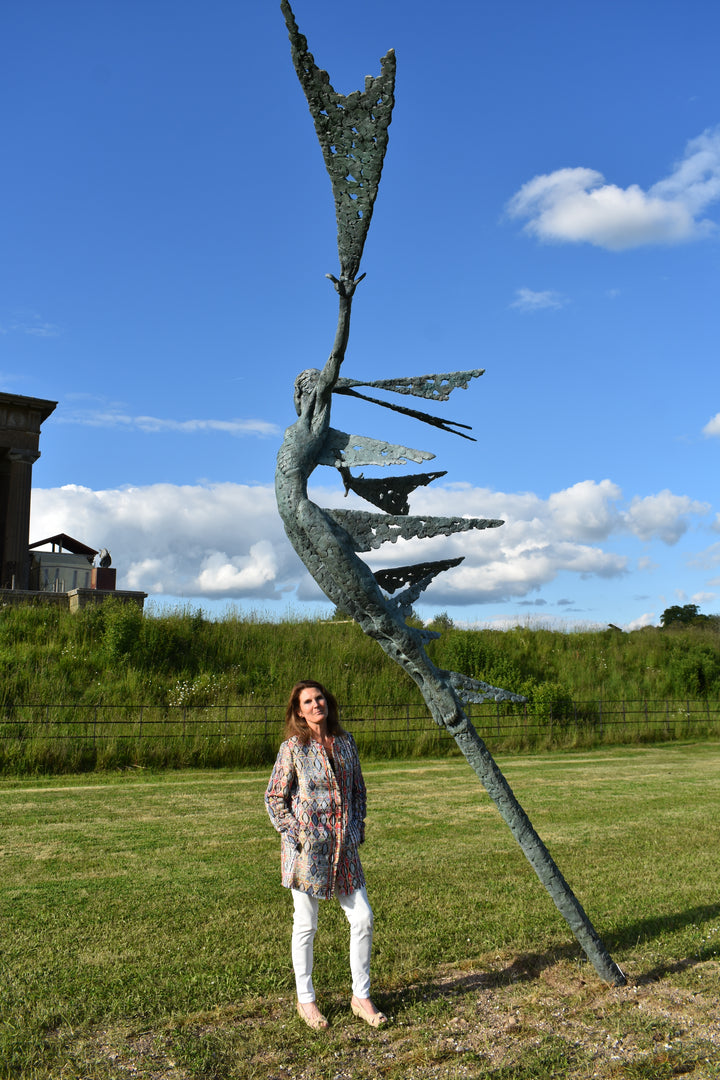 Monumental bronze sculpture of Theia, Greek goddess of light, portrayed in an elegant, elongated form with flowing limbs and wing-like extensions. Her textured surface and dynamic pose evoke movement and radiance, with light-catching shards projecting from her figure against open sky and landscape.