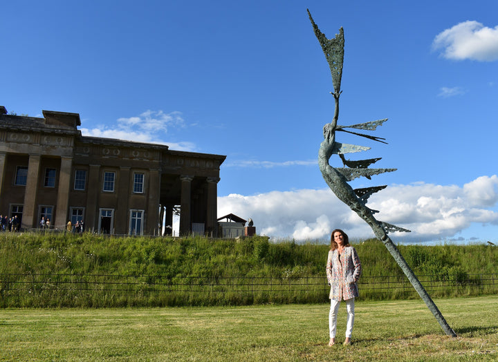 Monumental bronze sculpture of Theia, Greek goddess of light, portrayed in an elegant, elongated form with flowing limbs and wing-like extensions. Her textured surface and dynamic pose evoke movement and radiance, with light-catching shards projecting from her figure against open sky and landscape.