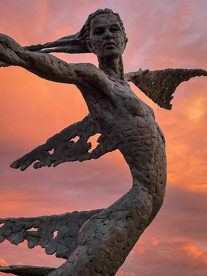 Monumental bronze sculpture of Theia, Greek goddess of light, portrayed in an elegant, elongated form with flowing limbs and wing-like extensions. Her textured surface and dynamic pose evoke movement and radiance, with light-catching shards projecting from her figure against open sky and landscape.