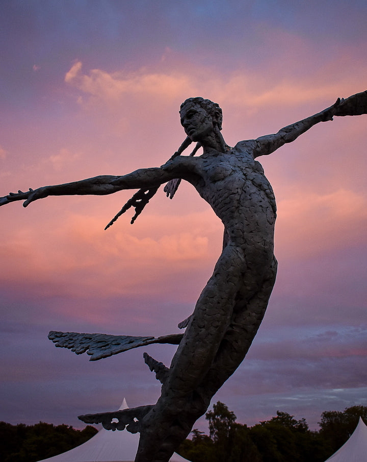 Monumental bronze sculpture of Theia, Greek goddess of light, portrayed in an elegant, elongated form with flowing limbs and wing-like extensions. Her textured surface and dynamic pose evoke movement and radiance, with light-catching shards projecting from her figure against open sky and landscape.