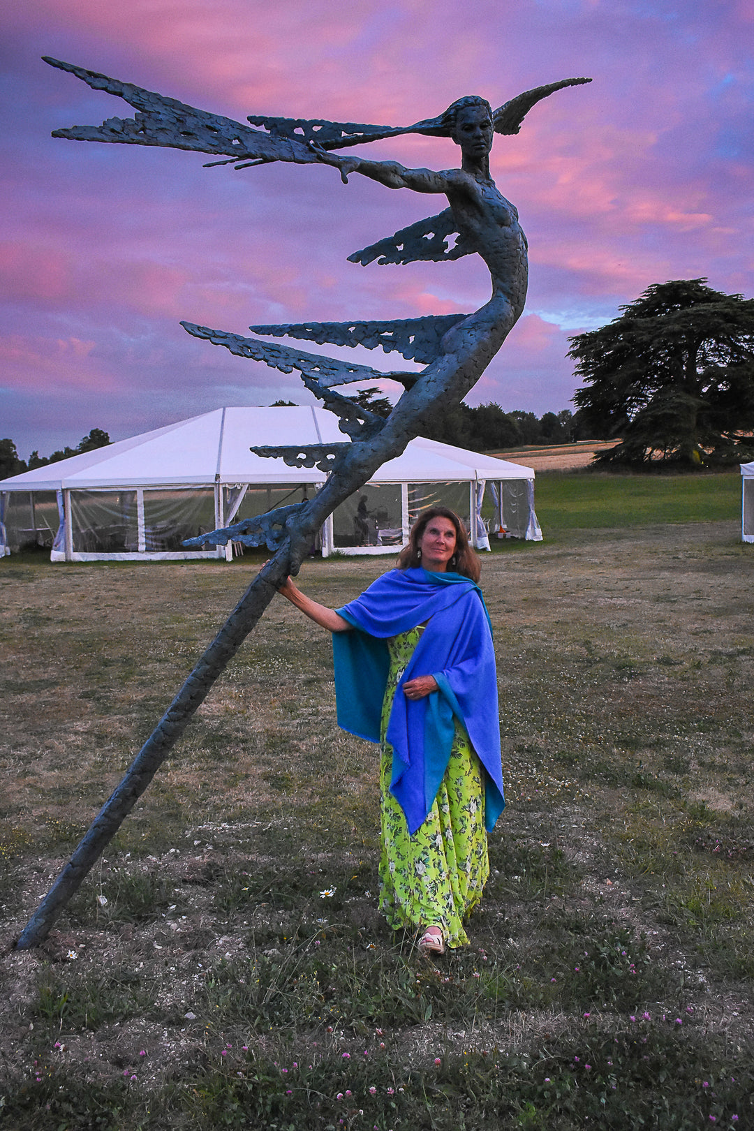 Monumental bronze sculpture of Theia, Greek goddess of light, portrayed in an elegant, elongated form with flowing limbs and wing-like extensions. Her textured surface and dynamic pose evoke movement and radiance, with light-catching shards projecting from her figure against open sky and landscape.