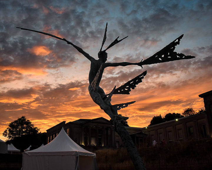Monumental bronze sculpture of Theia, Greek goddess of light, portrayed in an elegant, elongated form with flowing limbs and wing-like extensions. Her textured surface and dynamic pose evoke movement and radiance, with light-catching shards projecting from her figure against open sky and landscape.