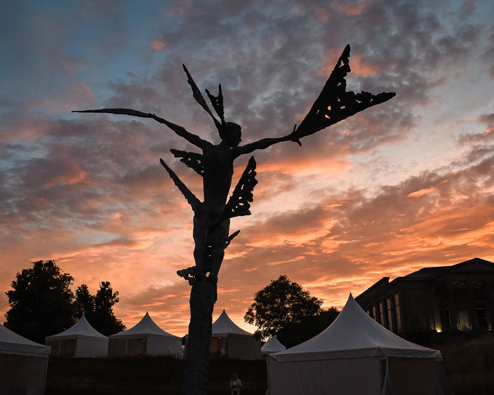 Monumental bronze sculpture of Theia, Greek goddess of light, portrayed in an elegant, elongated form with flowing limbs and wing-like extensions. Her textured surface and dynamic pose evoke movement and radiance, with light-catching shards projecting from her figure against open sky and landscape.