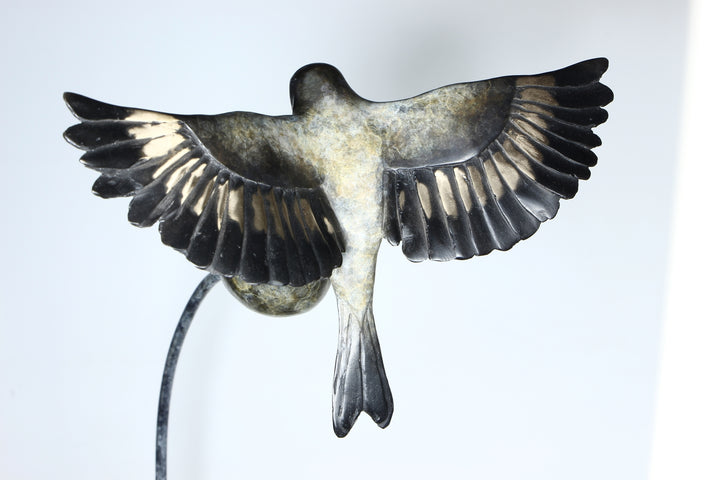 ‘Thistle Tweaker’ by Pat Bull, a limited-edition contemporary bronze wildlife sculpture depicting a goldfinch landing on a rounded teasel head with its wings fully outstretched. The bird features individually detailed bronze feathers, a distinctive reddish-brown face, dark wings with subtle bronze highlights and a forked tail. The teasel head is supported by a slender, sweeping curved steel stem rising from a rectangular bronze base, with dark grey, charcoal and muted green patina tones creating an elegant 