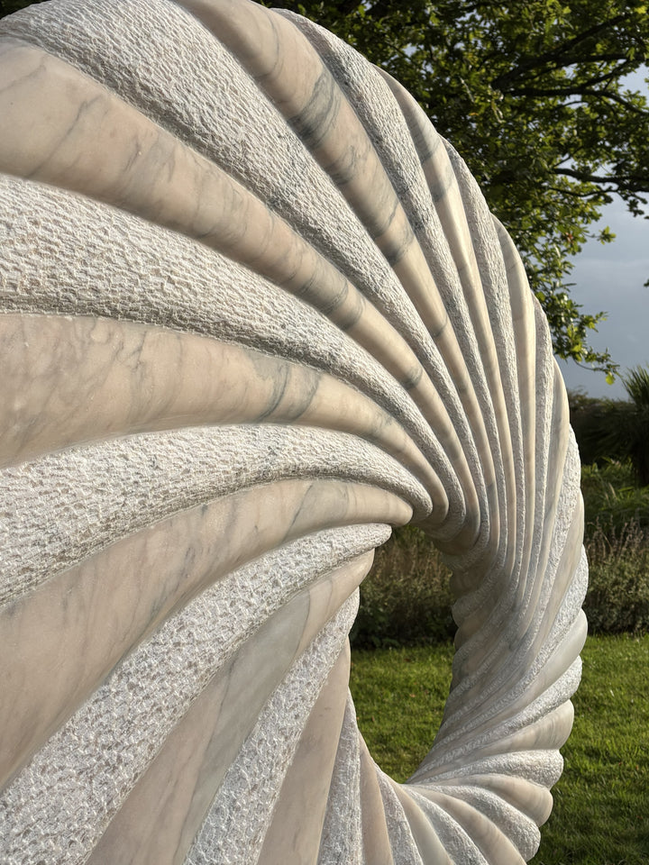 ‘Tidal II’ by Nicolas Moreton MRSS, a unique contemporary abstract stone sculpture carved from Portuguese Marble, featuring a large upright circular form with an open centre and deeply carved spiralling ridges. Alternating smooth, veined marble and finely textured stone surfaces create a twisting radial pattern around the central opening. Finished in natural shades of pale grey, white and charcoal-grey veining, the sculpture stands on a curved marble pedestal with a small arched opening, photographed in a l
