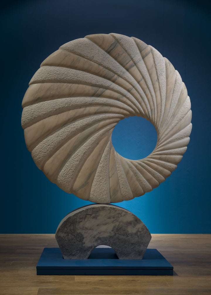 ‘Tidal II’ by Nicolas Moreton MRSS, a unique contemporary abstract stone sculpture carved from Portuguese Marble, featuring a large upright circular form with an open centre and deeply carved spiralling ridges. Alternating smooth, veined marble and finely textured stone surfaces create a twisting radial pattern around the central opening. Finished in natural shades of pale grey, white and charcoal-grey veining, the sculpture stands on a curved marble pedestal with a small arched opening, photographed in a l