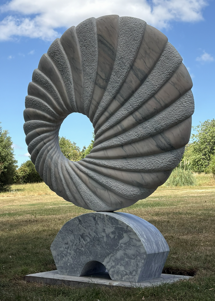‘Tidal II’ by Nicolas Moreton MRSS, a unique contemporary abstract stone sculpture carved from Portuguese Marble, featuring a large upright circular form with an open centre and deeply carved spiralling ridges. Alternating smooth, veined marble and finely textured stone surfaces create a twisting radial pattern around the central opening. Finished in natural shades of pale grey, white and charcoal-grey veining, the sculpture stands on a curved marble pedestal with a small arched opening, photographed in a l