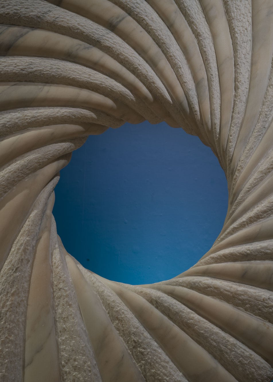 ‘Tidal II’ by Nicolas Moreton MRSS, a unique contemporary abstract stone sculpture carved from Portuguese Marble, featuring a large upright circular form with an open centre and deeply carved spiralling ridges. Alternating smooth, veined marble and finely textured stone surfaces create a twisting radial pattern around the central opening. Finished in natural shades of pale grey, white and charcoal-grey veining, the sculpture stands on a curved marble pedestal with a small arched opening, photographed in a l
