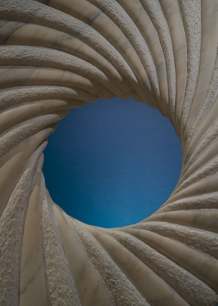 ‘Tidal II’ by Nicolas Moreton MRSS, a unique contemporary abstract stone sculpture carved from Portuguese Marble, featuring a large upright circular form with an open centre and deeply carved spiralling ridges. Alternating smooth, veined marble and finely textured stone surfaces create a twisting radial pattern around the central opening. Finished in natural shades of pale grey, white and charcoal-grey veining, the sculpture stands on a curved marble pedestal with a small arched opening, photographed in a l