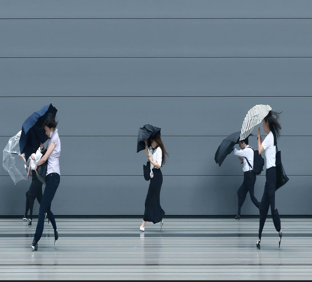 A stylised time-slice image of Tokyo commuters struggling against wind and rain during a summer storm. Each figure holds an umbrella as they move across a smooth, steel-grey background—some distorted mid-stride due to the artist’s high-speed video scan technique. The image captures both the uniformity and individuality of city life under pressure.
