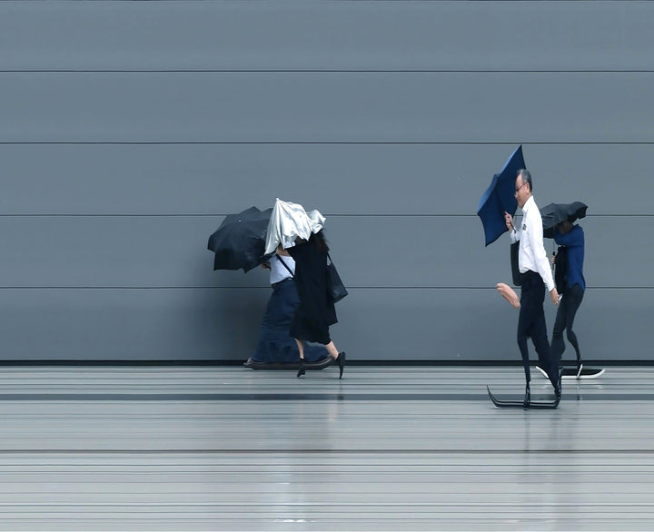 A stylised time-slice image of Tokyo commuters struggling against wind and rain during a summer storm. Each figure holds an umbrella as they move across a smooth, steel-grey background—some distorted mid-stride due to the artist’s high-speed video scan technique. The image captures both the uniformity and individuality of city life under pressure.